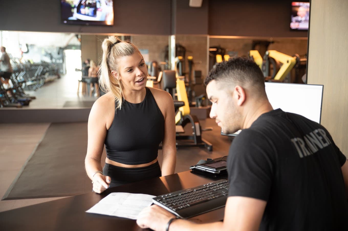 A man and woman talking at a desk in a gym.