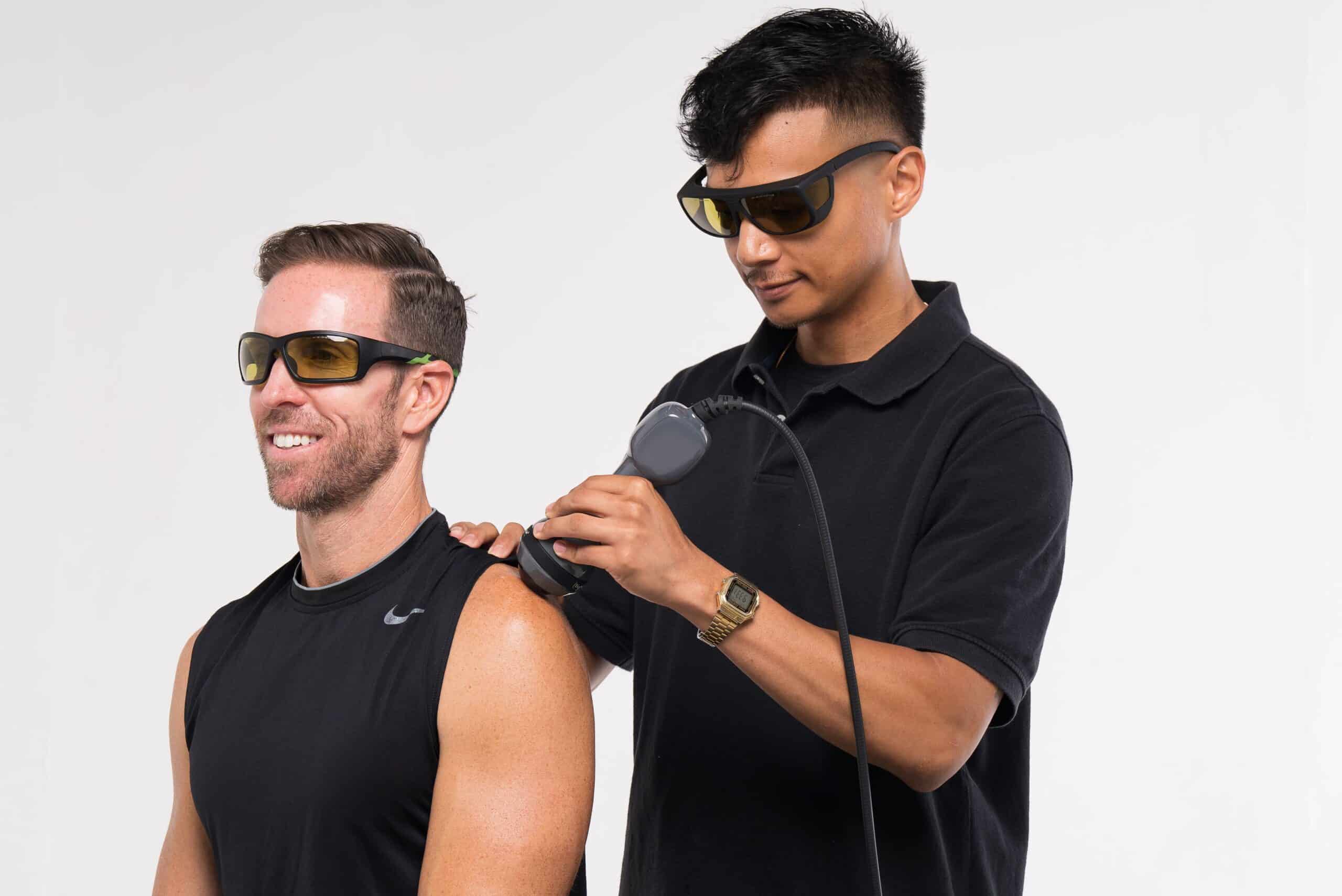 A man is getting his arm shaved by a man wearing sunglasses.