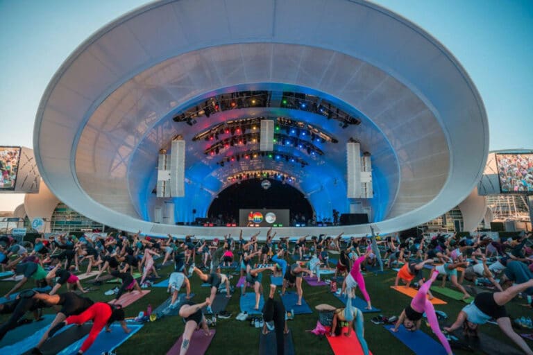 Outdoor yoga session at a concert venue with participants on colorful mats and a stage in the background.