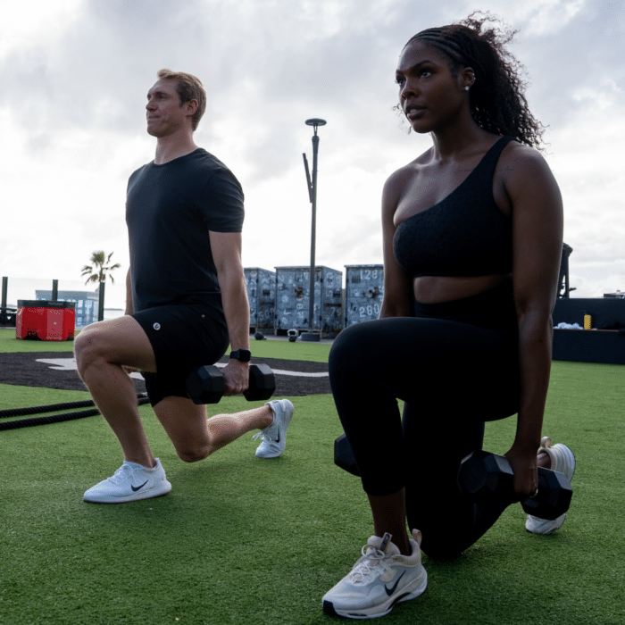 Two people performing lunges outdoors with dumbbells.