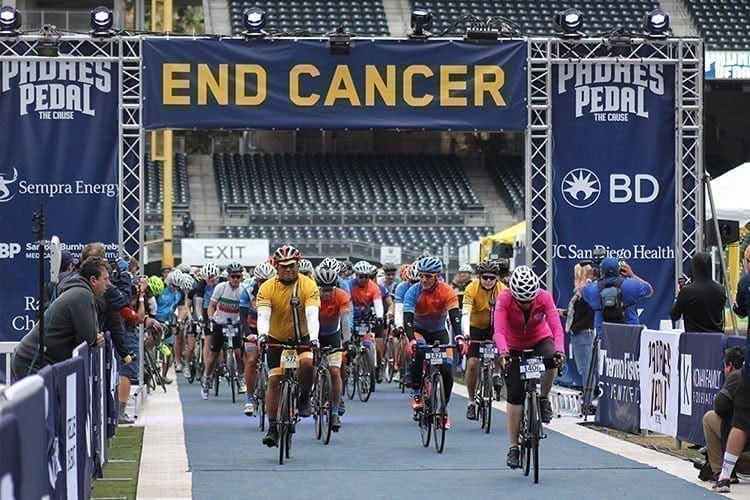 Cyclists riding through a stadium under a banner that reads "end cancer" at a charity event sponsored by multiple companies.
