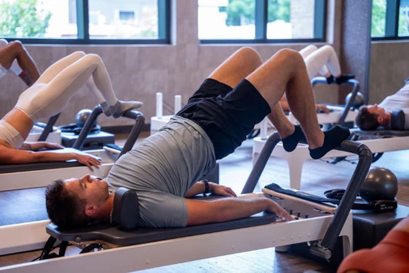 People in a fitness studio are exercising on reformer machines, with legs raised and gripping handles, focusing on core strength and stability.