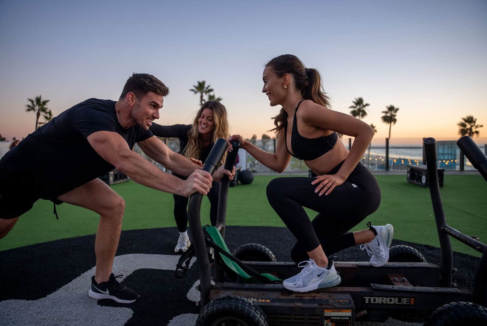 Three people exercising outdoors at sunset, two women pushing a sled and a man helping them, with palm trees and ocean in the background.