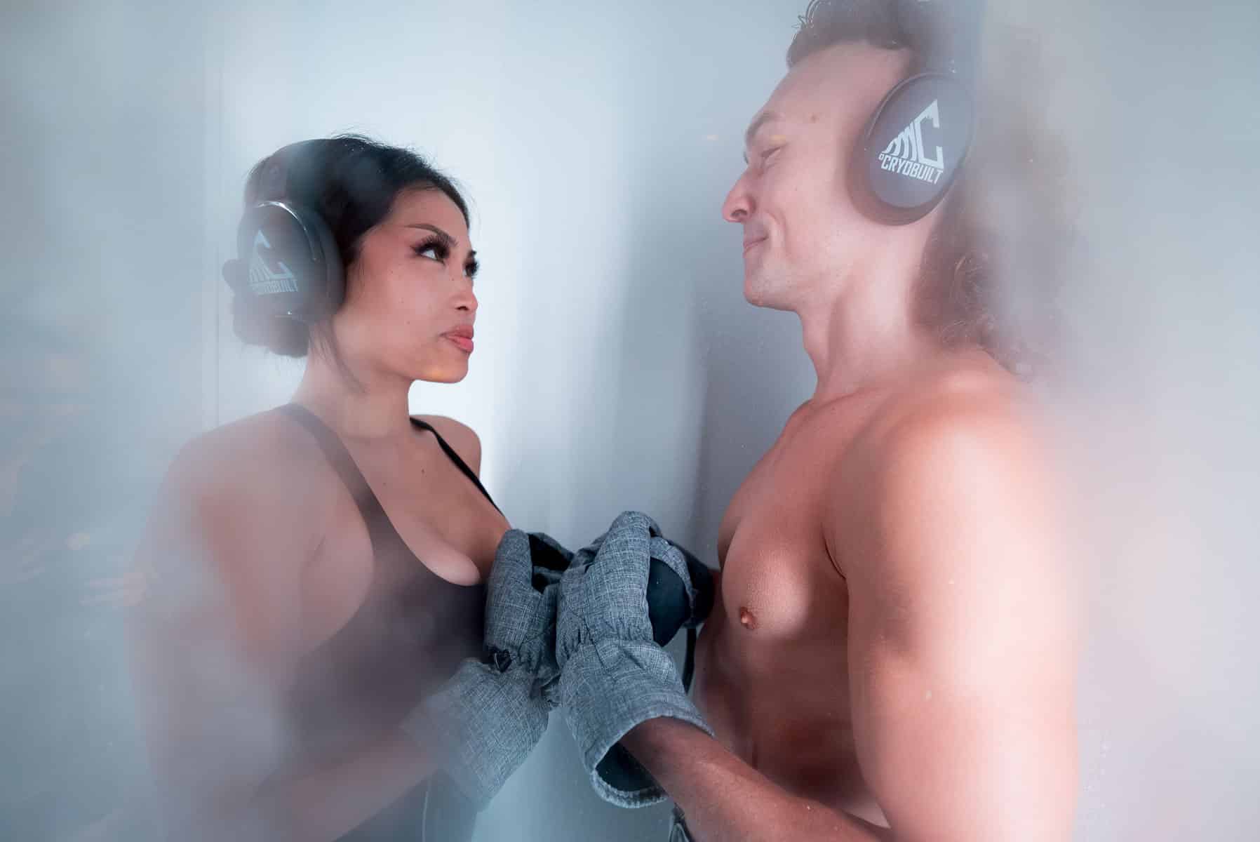 Two people in a cold chamber wearing headphones and gloves face each other with hands clasped, surrounded by mist.