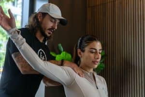 A man assists a woman in a stretching exercise. The woman has one arm raised and the man supports her shoulder. They are indoors with a wooden panel background and a plant visible.