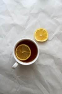 A cup of tea with a lemon slice on a caramelized white surface. There is a separate lemon slice nearby.