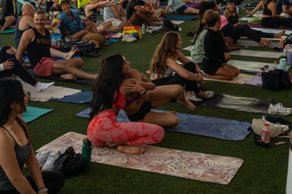 People sitting on yoga mats, some wearing athletic clothing. Two individuals seated in the foreground embrace. The setting appears to be a large group event on grass.