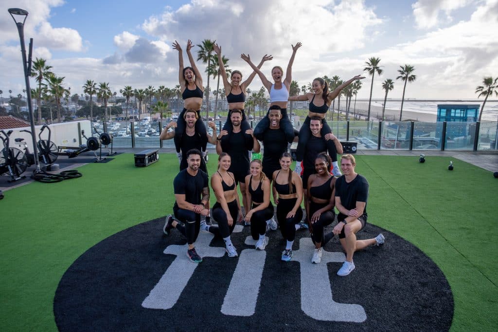 A group of people in athletic wear posing outdoors on a grassy area with exercise equipment, palm trees, and a cloudy sky in the background.