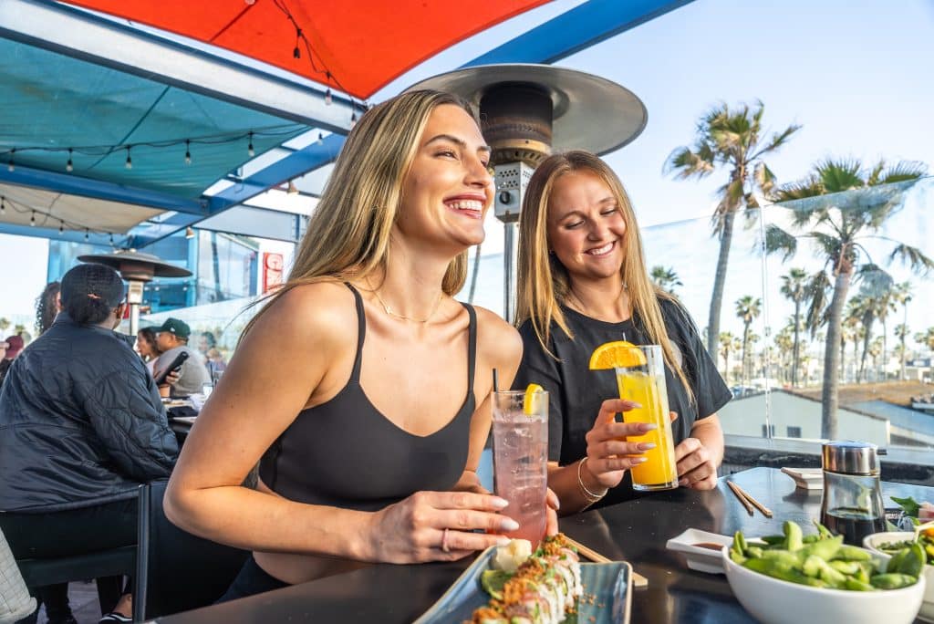 Two women at an outdoor cafe table, smiling and holding drinks. Plates of food are visible in front of them. Palm trees and a clear sky are in the background.